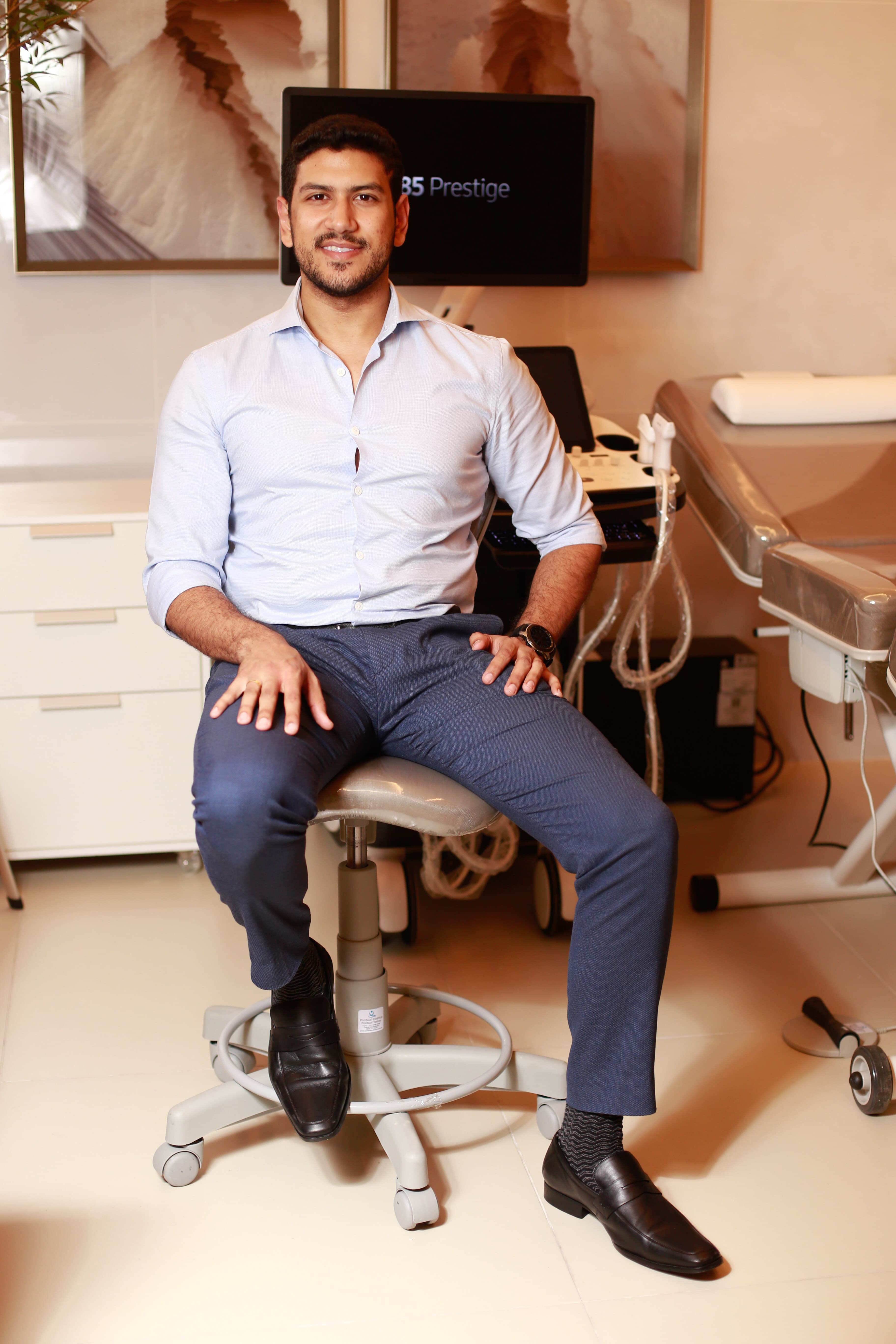 Smiling bearded man in a blue shirt sits on a stool in a medical office.